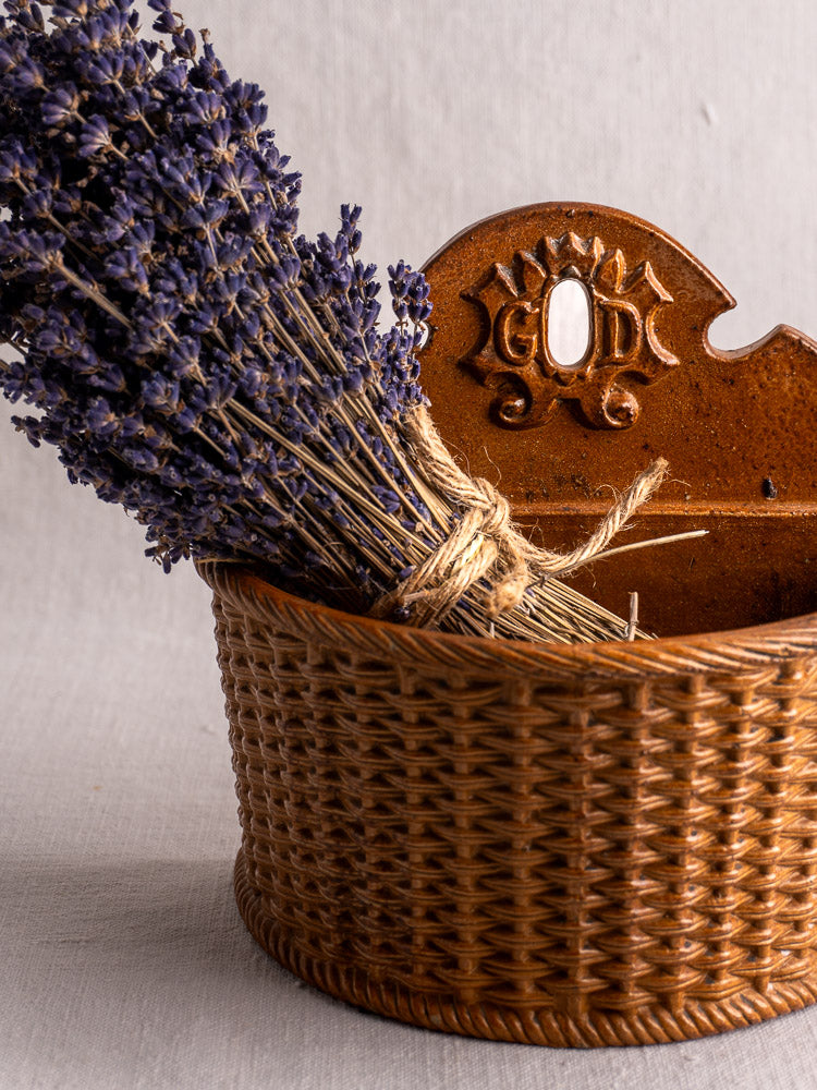 Antique French salt-glazed stoneware salt box beside dried lavender in a brown woven basket with “G O D” sunburst backplate.