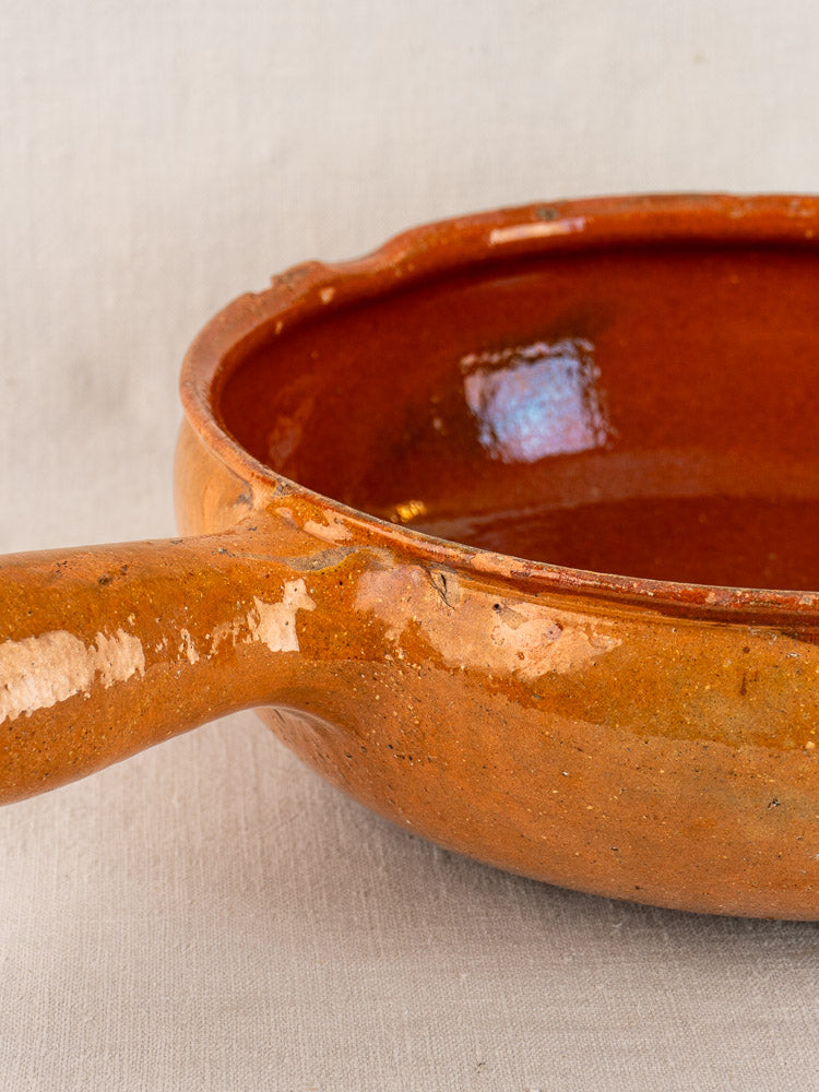 Antique French Vallauris terracotta poêlon with orange-brown glaze and handle, set on a light linen cloth.