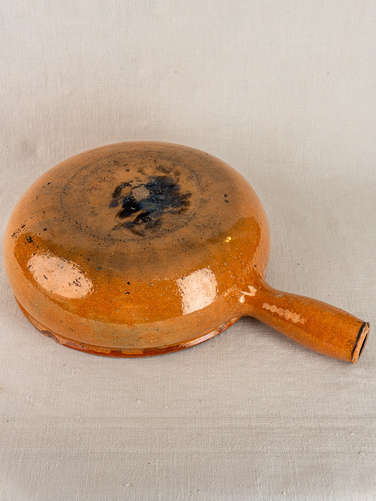 Early 20th-century Vallauris glazed terracotta poêlon upside down, glossy orange-brown surface on light fabric.