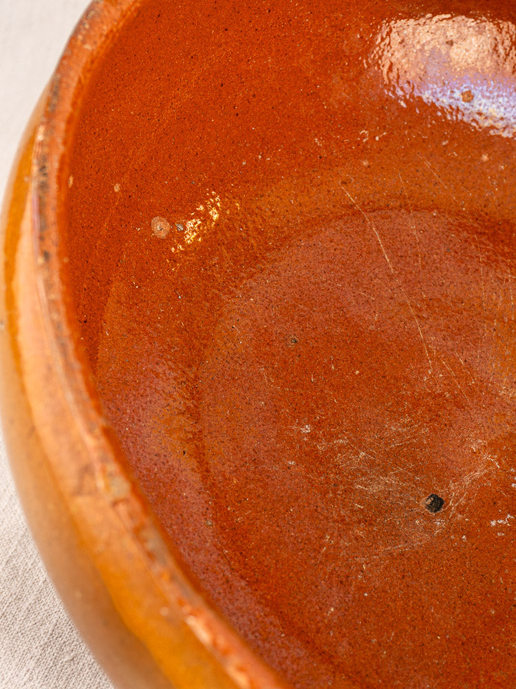 Antique French Vallauris glazed terracotta poêlon bowl, reddish-brown interior with scratches and chip, on light fabric background.