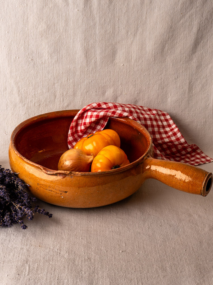 Antique glazed Vallauris terracotta poêlon with yellow tomatoes, onion, and checkered cloth; dried lavender alongside. Early 20th century.