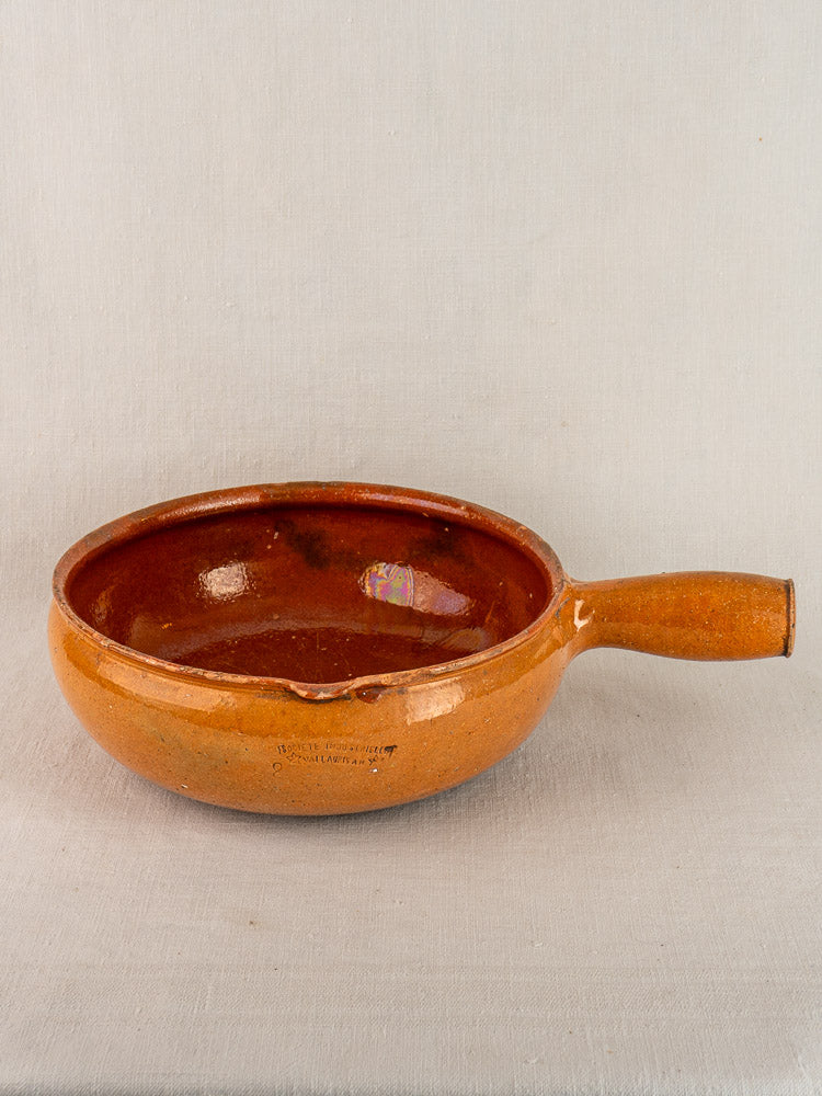 Antique French Vallauris poêlon in glazed terracotta with right handle and glossy interior on light fabric background.