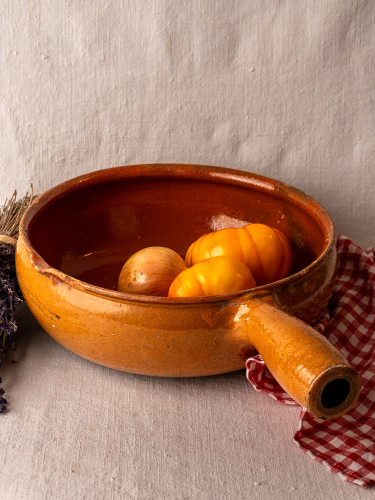 Antique French Vallauris glazed terracotta poêlon with handle, holding heirloom tomatoes and onion on red checked cloth, dried lavender nearby.