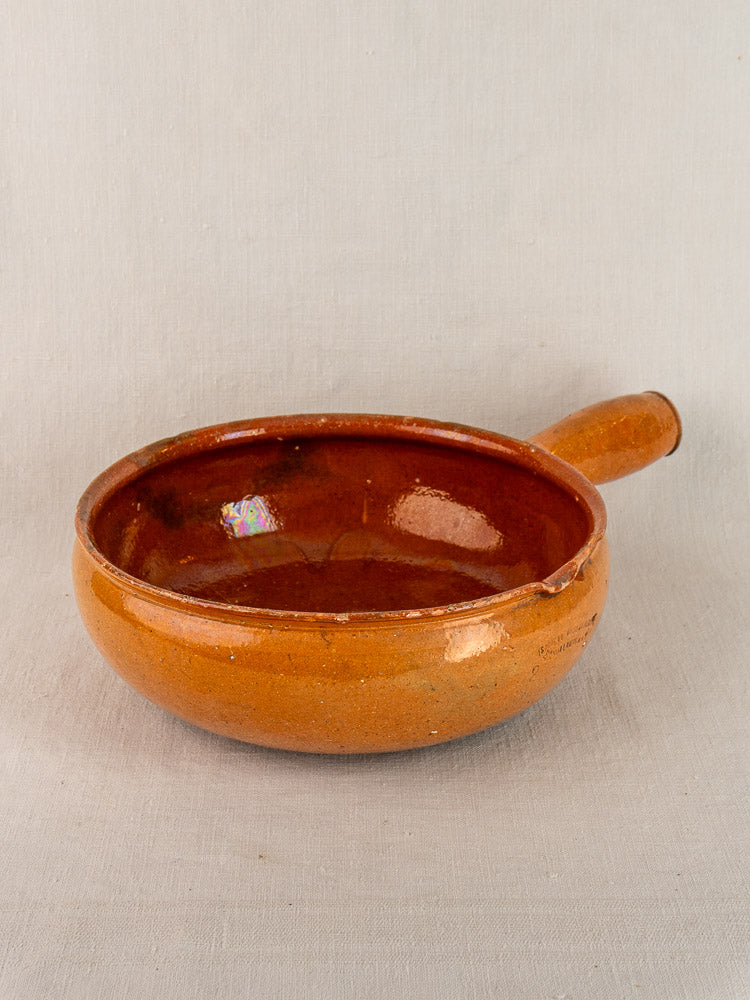 Early 20th century Vallauris French poêlon, round glazed terracotta with a single short handle, on a plain light background.