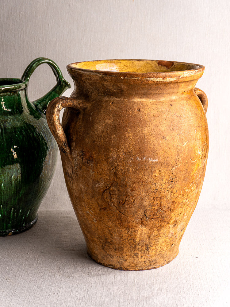Antique French terracotta storage jar with yellow glazed interior beside a green glazed jug, neutral background.