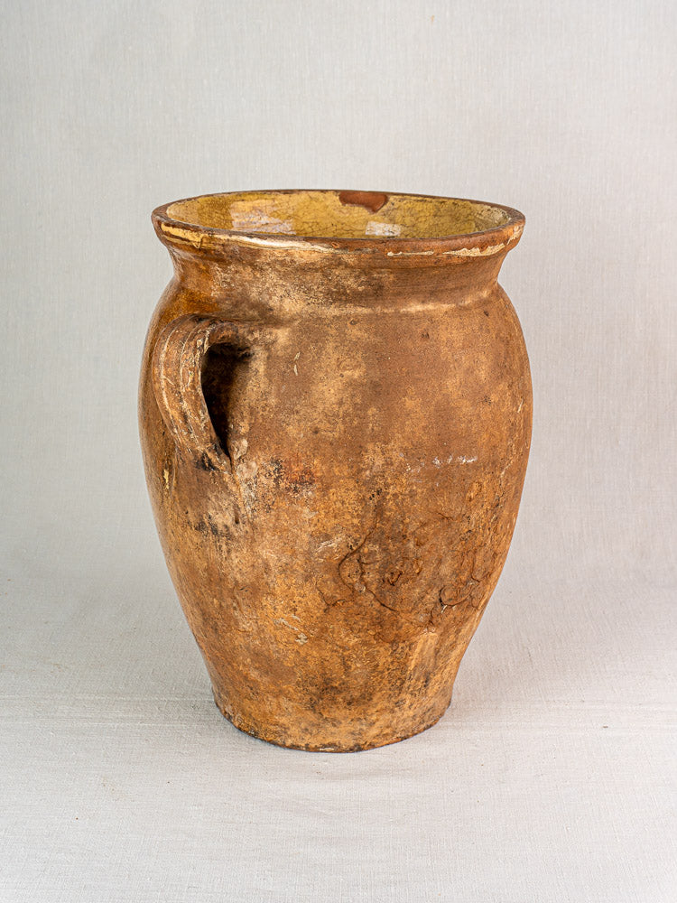 Antique French terracotta storage jar with a single handle and yellow glazed interior on textured surface.