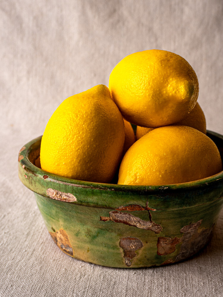 Antique French terracotta tian bowl with green and yellow glaze, chipping, on textured surface.