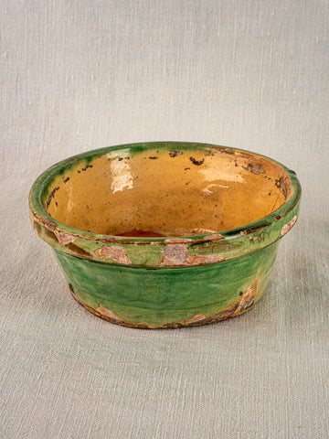 Antique French terracotta tian bowl with green and yellow glaze, chipping, on textured surface.