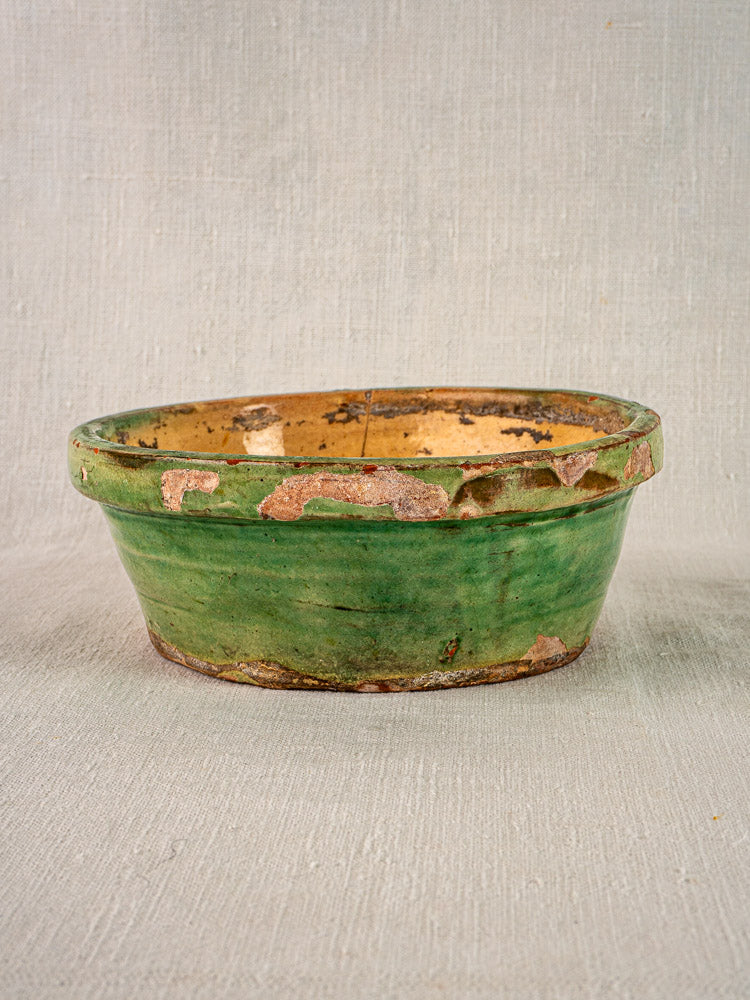 Antique French tian bowl with worn green and yellow glaze, exposed terracotta, on textured surface.