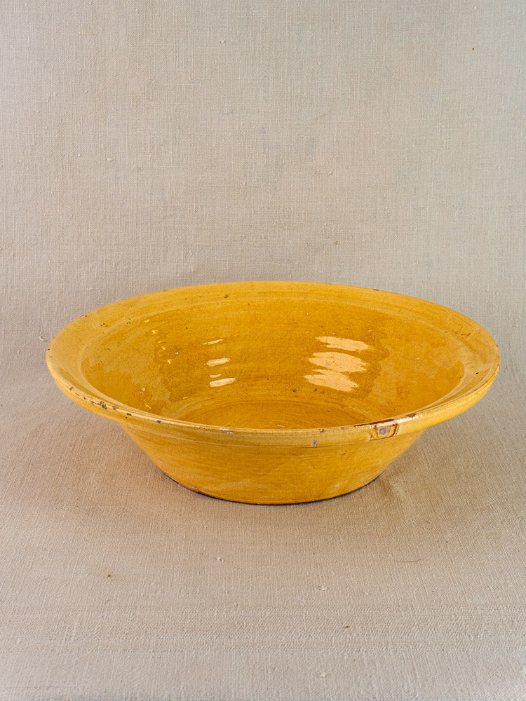 Antique French yellow glazed terracotta tian bowl, wide and shallow, on a neutral textured surface.