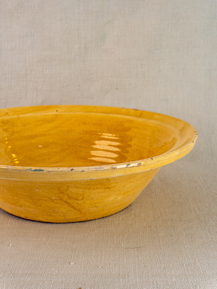 Antique French yellow glazed tian bowl with chipped rim on a beige textured surface.