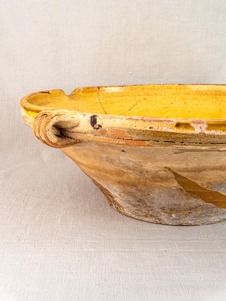 Rustic shallow ceramic bowl with saffron-yellow glazed rim and handle, unglazed weathered base, on light fabric background.