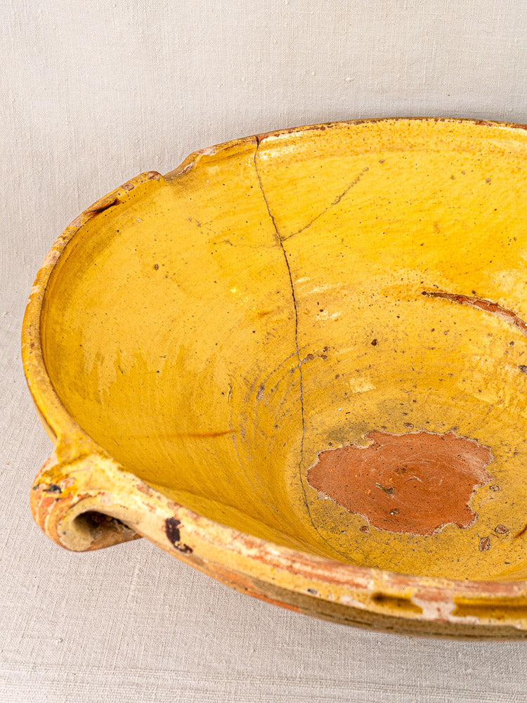 Saffron-yellow glazed Provençal Tian bowl with handle, rustic cracks, chips, and reddish-brown interior patch on neutral fabric.