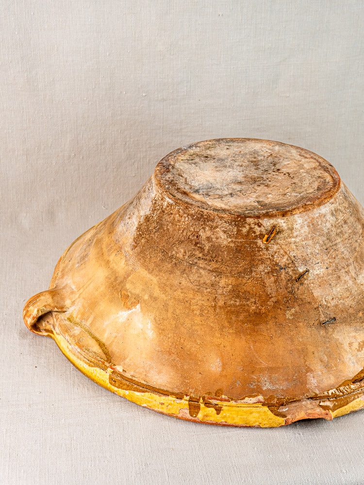 Large rustic Provençal yellow tian bowl with weathered terracotta and small handle on neutral fabric.