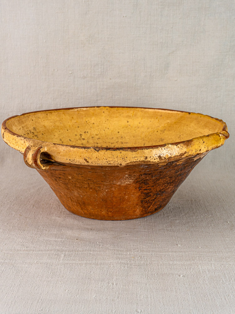 Rustic Provençal yellow tian bowl with wide rim, twin handles, yellow glazed interior, brown exterior on fabric background.
