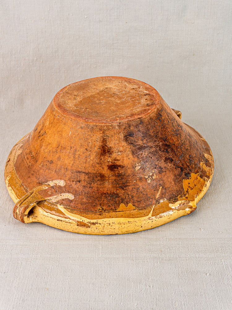 Antique Provençal yellow tian bowl with side handles, saffron glaze, and worn, chipped edges on light fabric.