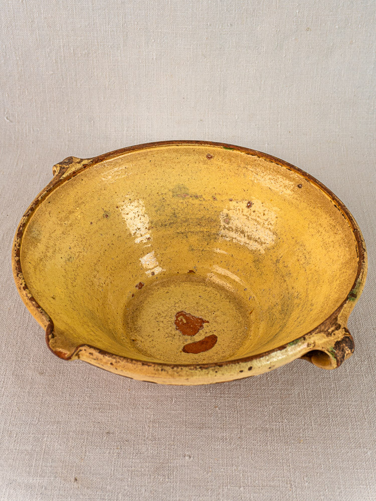 Antique Provençal yellow tian bowl with rustic glaze, two handles, on a light beige fabric background.