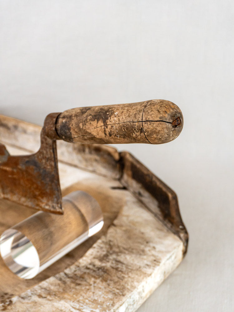 Antique French wood and iron tobacco cutter with worn handle, on weathered wood next to glass cylinder.