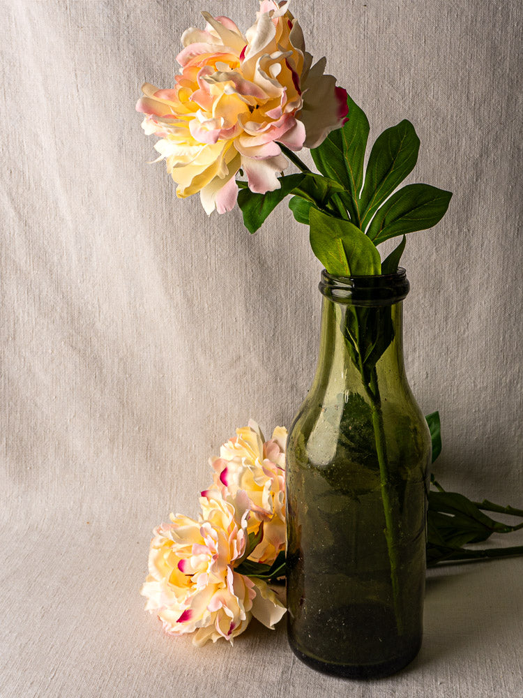 Antique French truffle bottle in hand-blown olive glass with peonies, beige fabric backdrop.