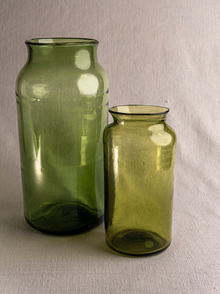 Pair of antique French olive green glass preserve jars, hand blown, on a light gray textured surface with soft shadows.