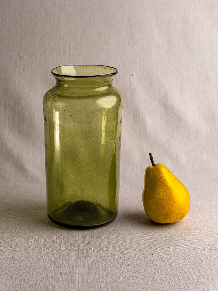 Antique French olive green glass preserve jar, late 18th century, with yellow pear on textured fabric background.