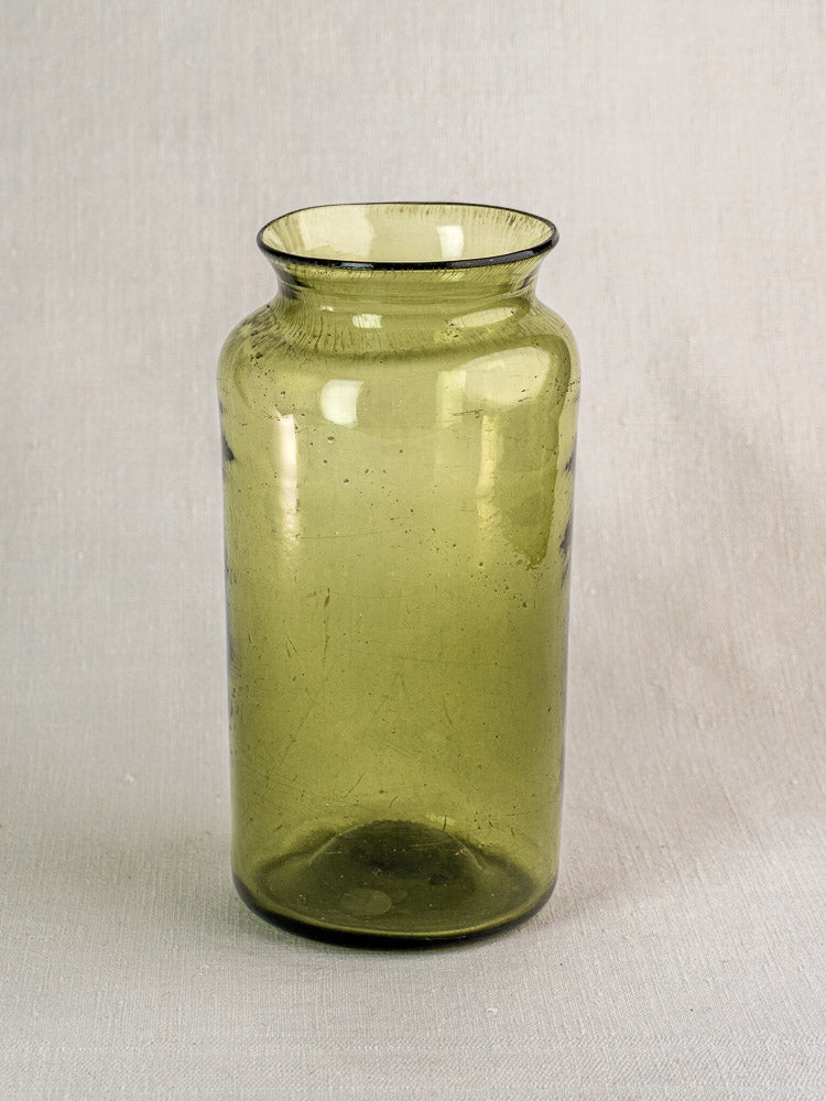 Antique French olive green glass preserve jar, tall with wide mouth and flared rim, on light gray textured surface.