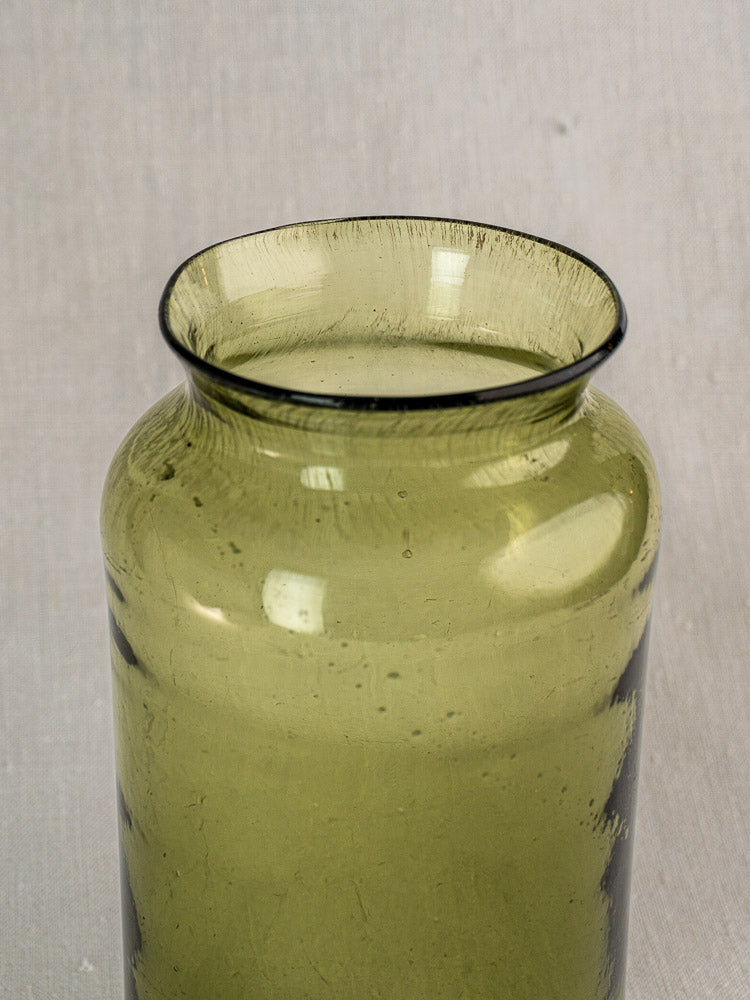 Antique French preserve jar in hand-blown olive green glass, wide opening and flared rim on neutral fabric background.