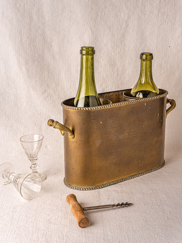 Antique French brass double champagne cooler with green wine bottles, wine glasses, and corkscrew on white surface.