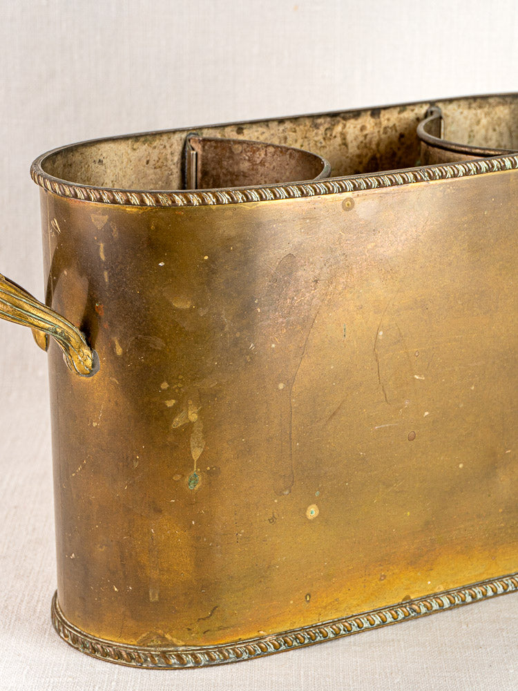 French antique brass double champagne cooler with rope edging, side handle, and natural tarnish on light fabric.