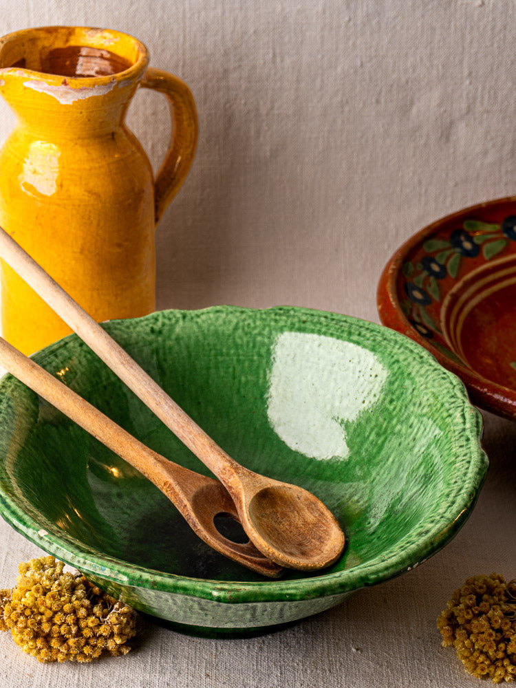 French Vallauris bowl with green glaze, wooden spoons, yellow ceramic pitcher, red bowl, and dried flowers on beige fabric.