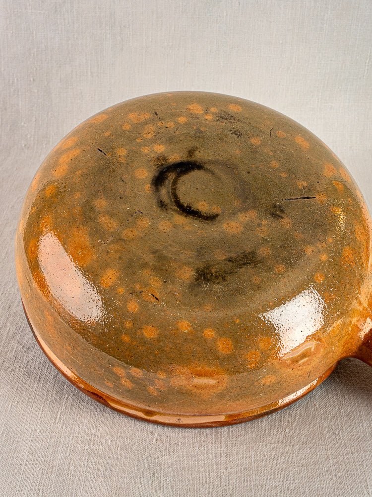 Antique French terracotta cooking pan with orange-brown speckled glaze and central dark crescent, displayed upside down on fabric.