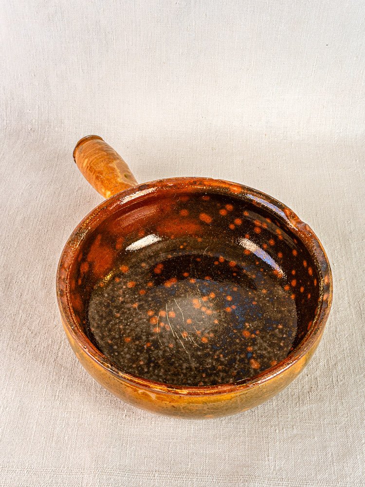 Antique French Vallauris Jaspé terracotta poêlon with handle and rustic brown speckled glaze on light textured surface.