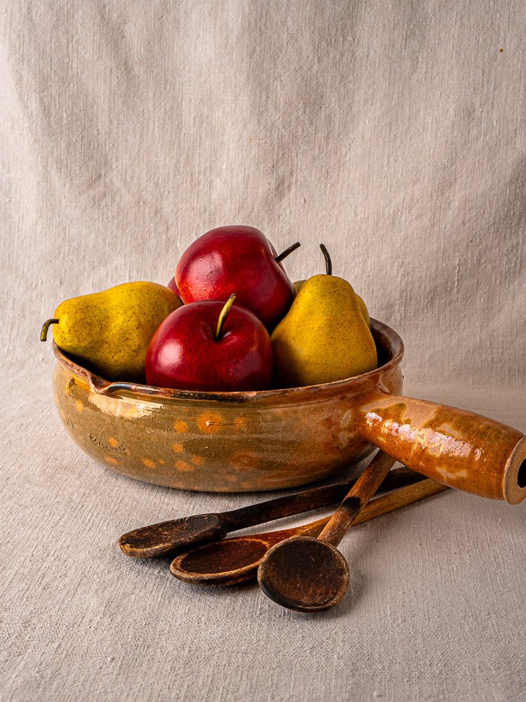 Antique French Vallauris Jaspé terracotta poêlon with apples, pears, and wooden spoons on cloth.