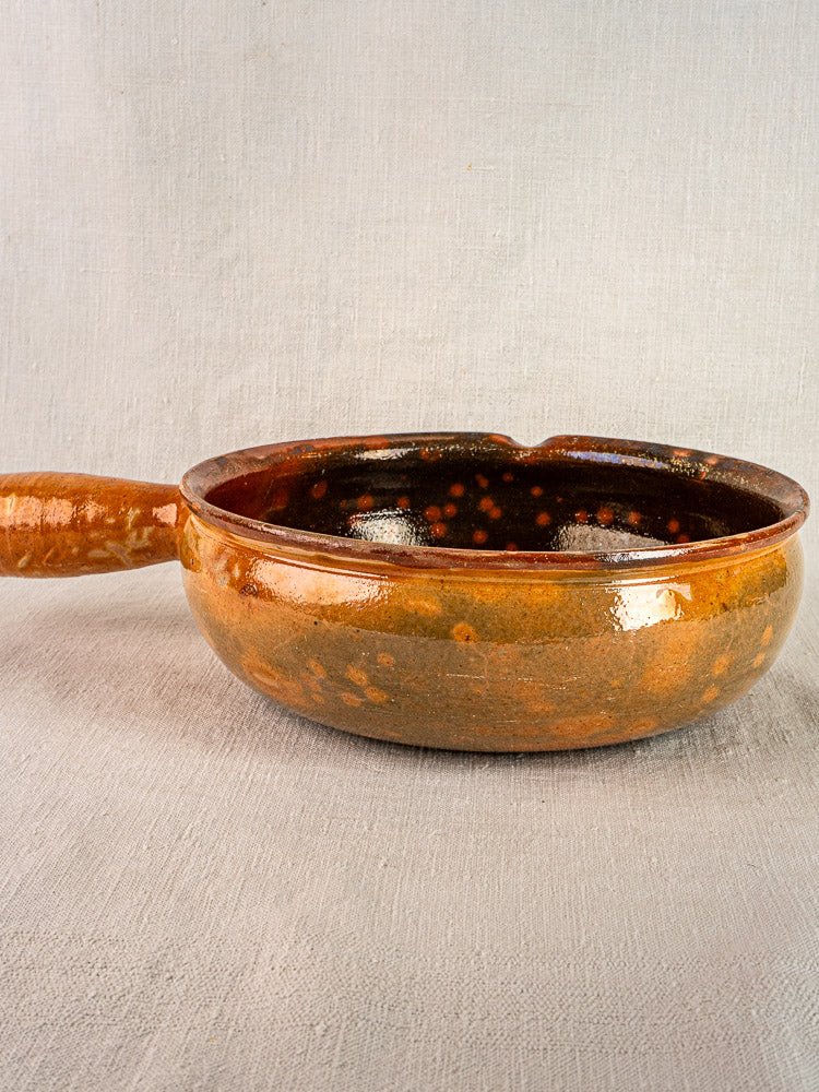 Antique French terracotta Vallauris Jaspé poêlon with speckled brown glaze and pouring spout on light fabric.