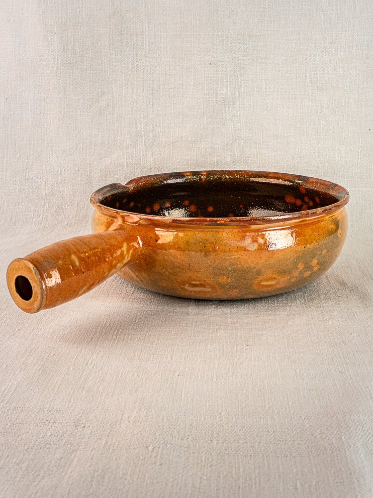 Antique French Vallauris Jaspé Poêlon terracotta pan with brown glaze and single handle on light fabric.