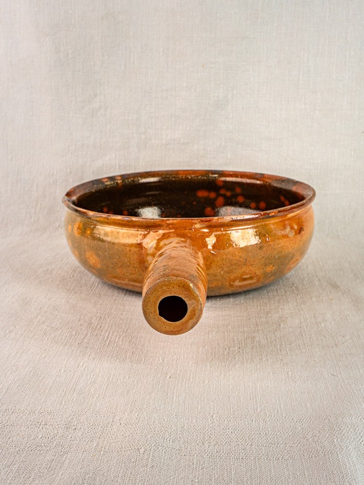 Round brown ceramic Vallauris Jaspé Poêlon terracotta pan with spout on textured surface, light gray backdrop.