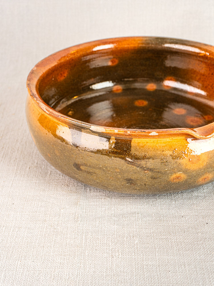Antique French Vallauris terracotta poêlon, glossy brown glaze with orange flecks, on light gray textured surface.