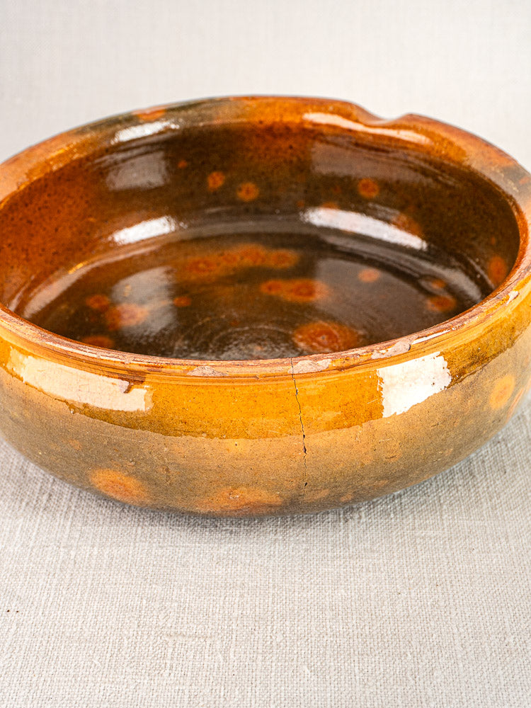 Antique French Vallauris poêlon, round glazed ceramic with brown-orange mottled finish, minor crack visible, on light fabric.