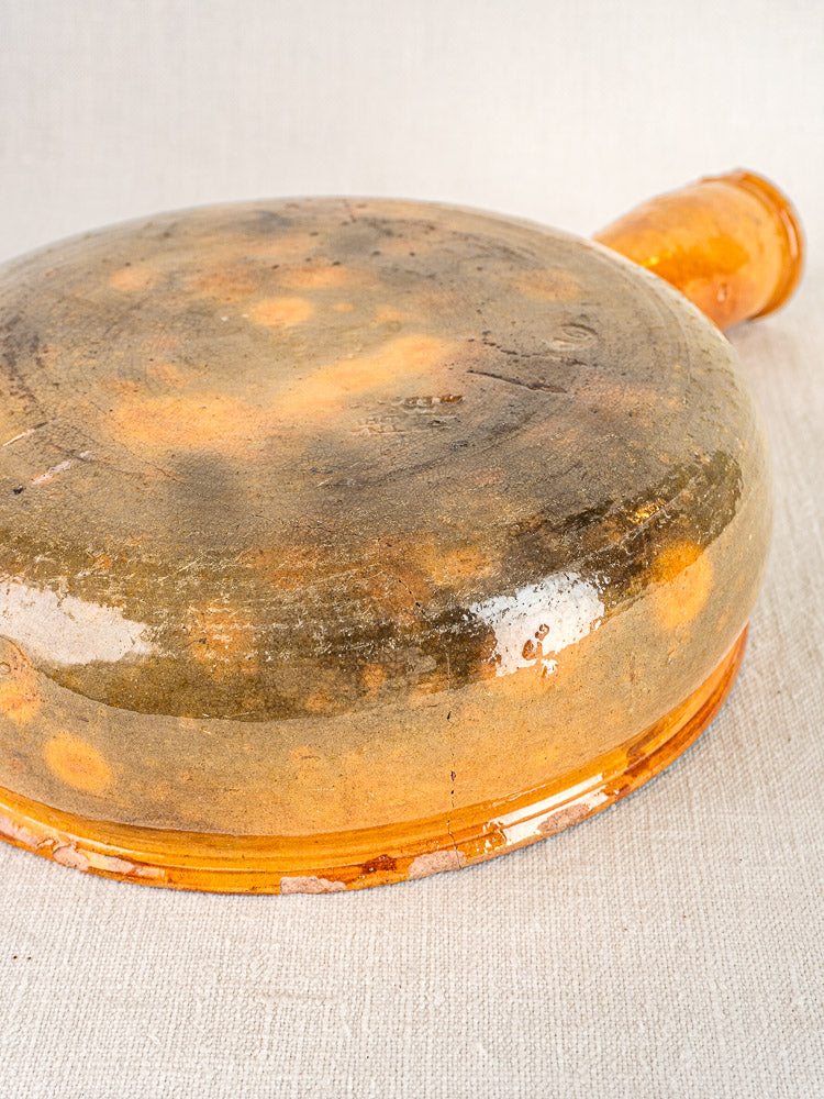 Antique French Vallauris terracotta poêlon with brown-orange glaze, flat base, and lateral spout on fabric.