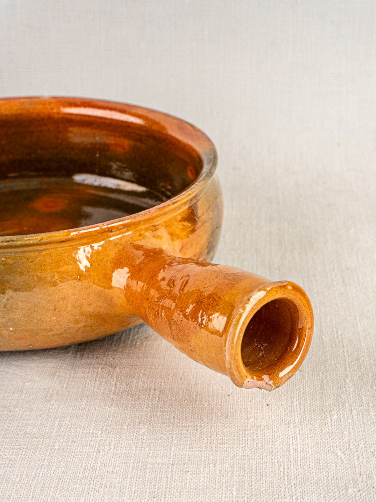 Antique French Vallauris terracotta poêlon with rich brown glaze and short spout on textured surface.