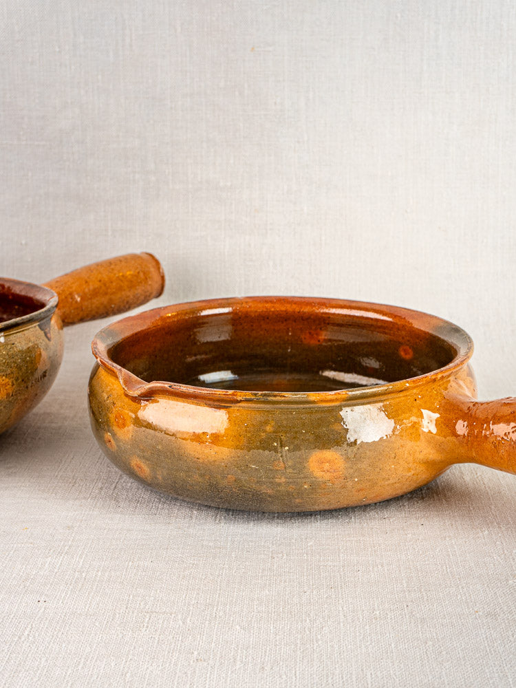 Antique French Vallauris terracotta poêlons with handles, glossy glaze, on light textured surface.