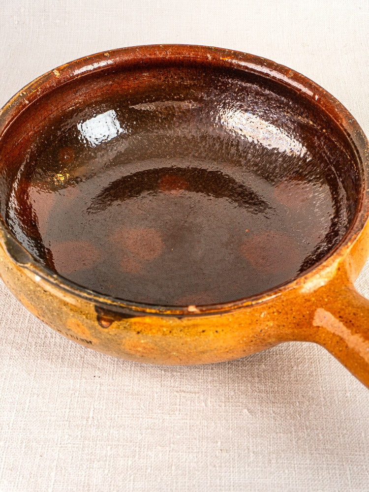 Antique French Vallauris poêlon, brown glazed ceramic with handle, on light fabric surface.