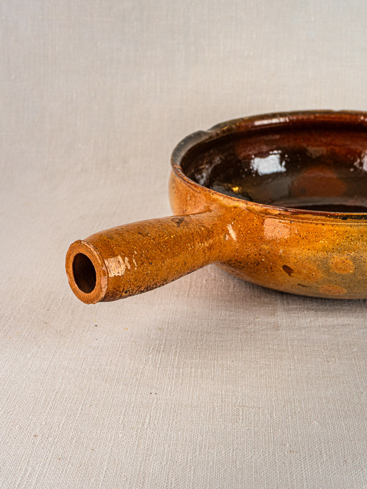 Antique French Vallauris brown glazed ceramic poêlon with hollow handle on light fabric background.
