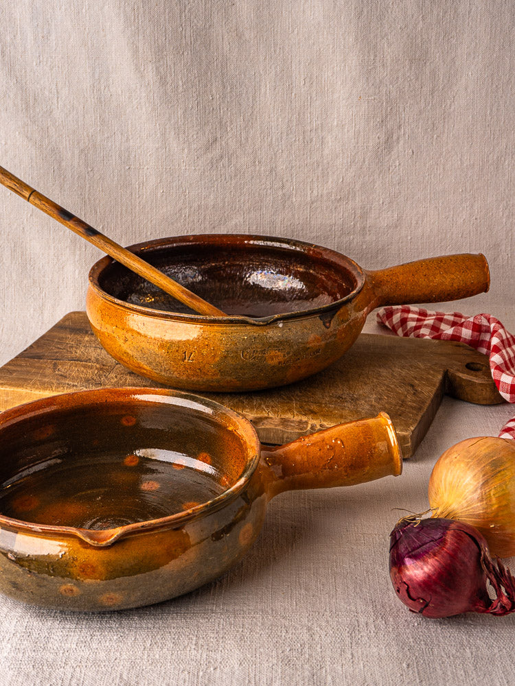 Pair of antique French Vallauris terracotta poêlons on wood with spoon and onions.