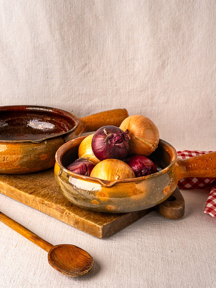 Pair of antique French Vallauris terracotta poêlons on wooden board with onions and spoon.