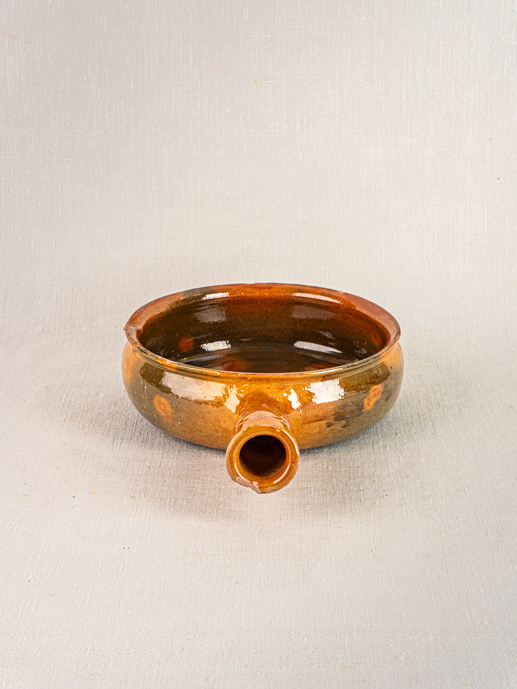 Antique French Vallauris terracotta poêlon, round shallow brown ceramic with short spout, on neutral textured background.