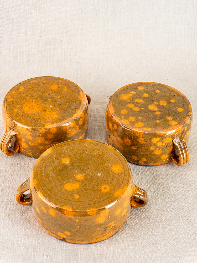 Set of three vintage French Vallauris bowls in mottled brown glazed earthenware with yellow spots and side handles, on beige fabric background.