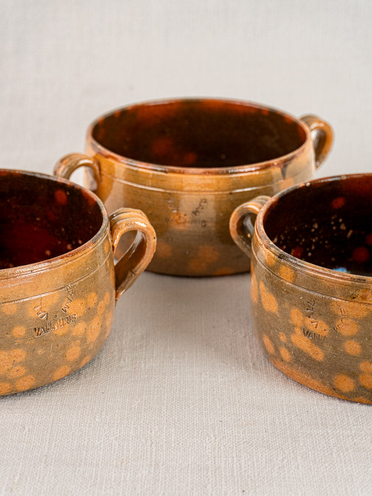 Set of three vintage French Vallauris earthenware bowls with orange speckled glaze and handles on light fabric.