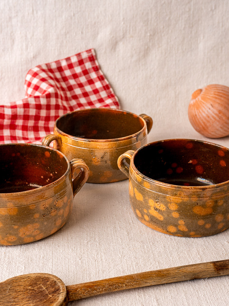 Set of three vintage French Vallauris ceramic bowls with mottled brown glaze and side handles on textured fabric.