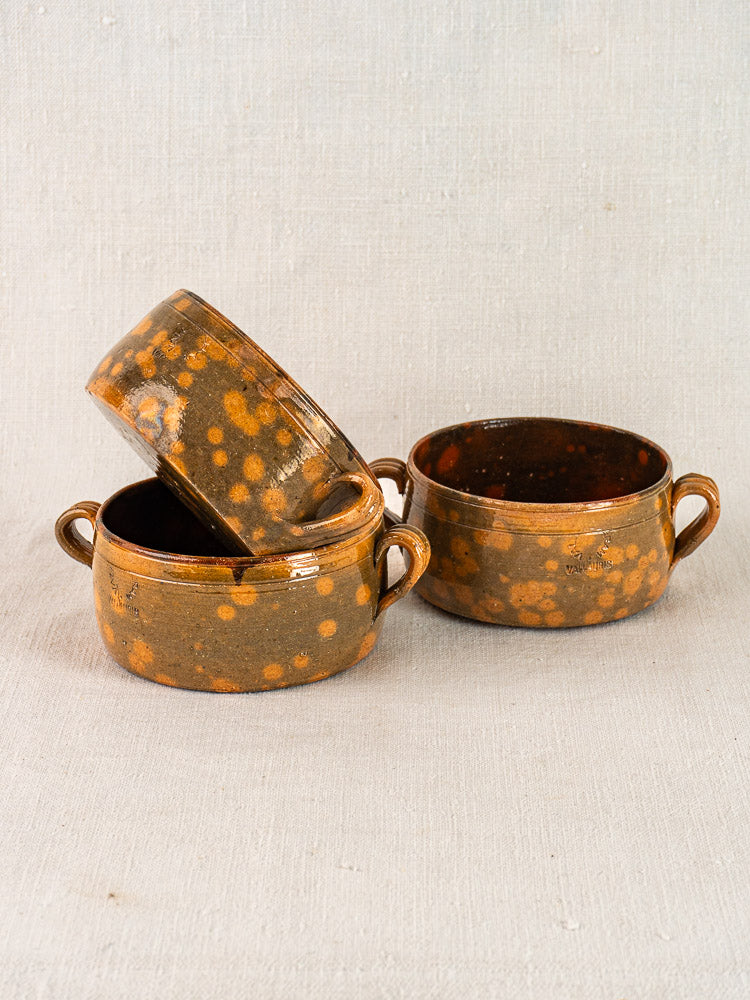 Set of three vintage French Vallauris ceramic bowls with mottled brown glaze and side handles on textured fabric.